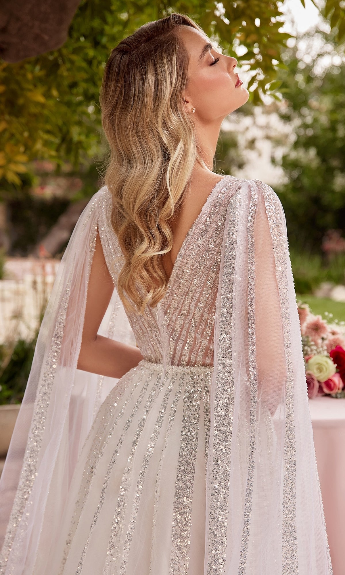 A woman with long wavy blonde hair wears the Ladivine CD852W Drape-Sleeve White Formal Dress, a sparkly backless gown with silver stripes, while standing outdoors near a flower-adorned table.